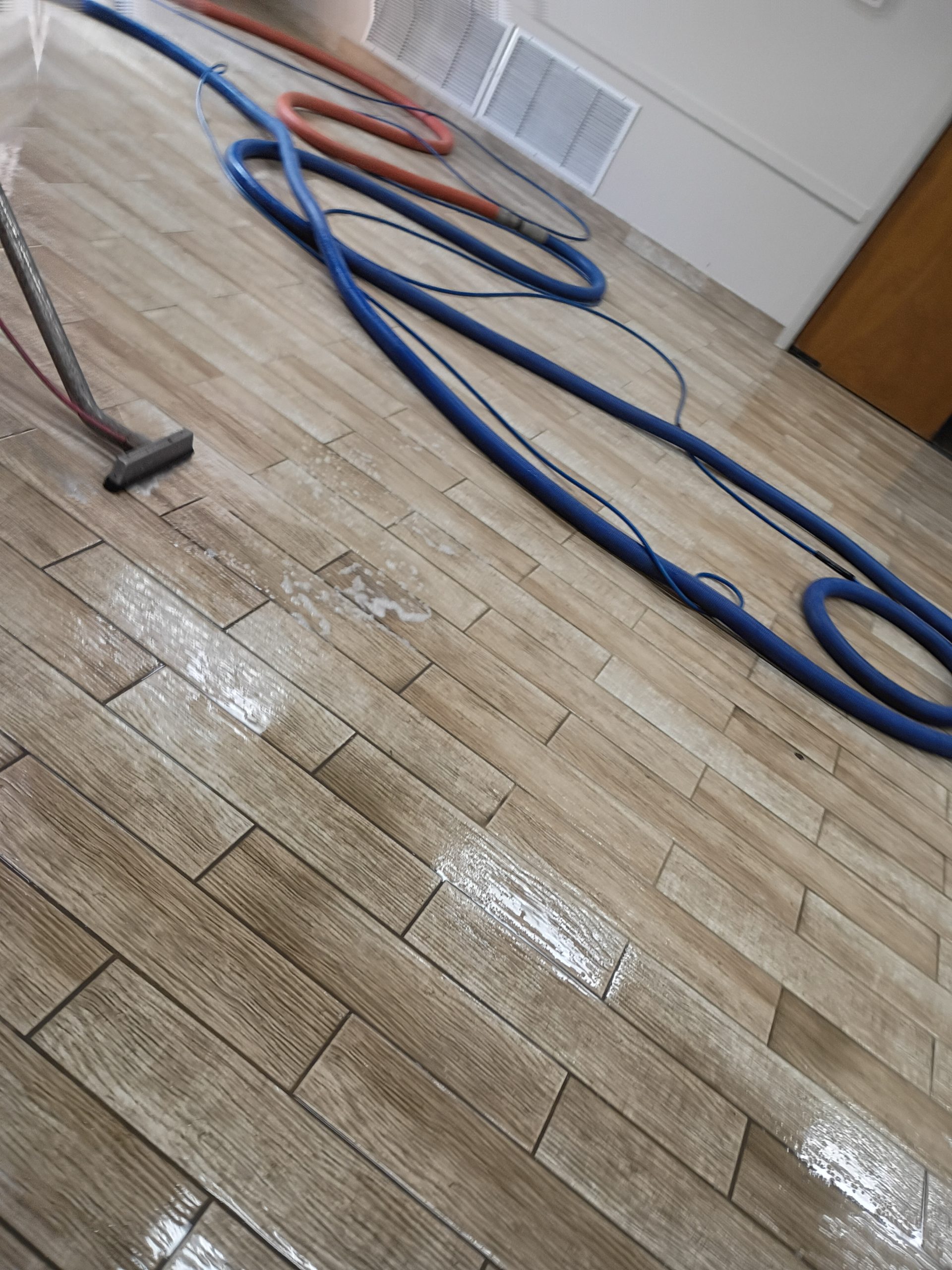 Tile cleaning equipment hoses coiled on a wet wood-look plank floor, with a cleaning wand visible at the edge of the frame.