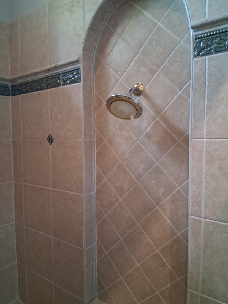 A shower stall with an arched doorway opening, covered in tan diamond-pattern tile bordered by a decorative dark blue and gold mosaic trim near the top, with a gold-toned showerhead mounted on the back wall and a small diamond-shaped tile accent on the side wall.