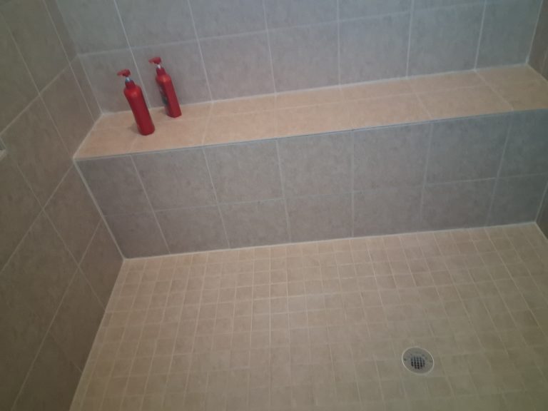 Shower stall with grey tile walls, a tiled bench holding two red soap bottles, and a beige mosaic tile floor with a drain.