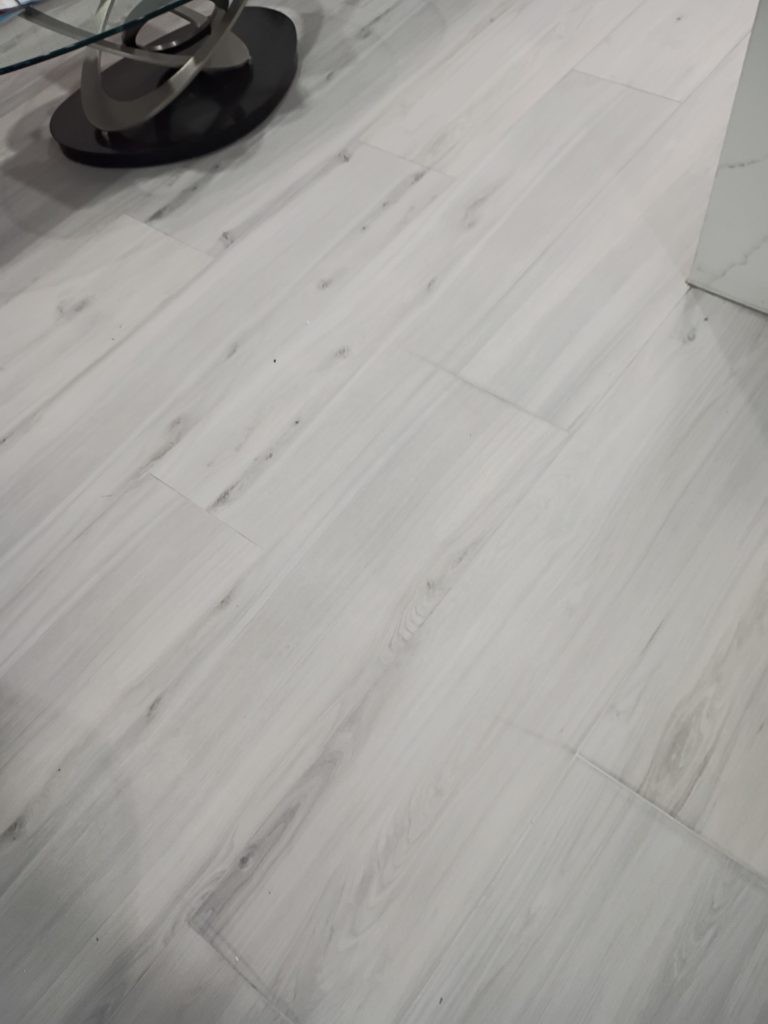 White and grey wood-look plank flooring in a living room, with a modern glass and metal coffee table sculpture visible in the corner.