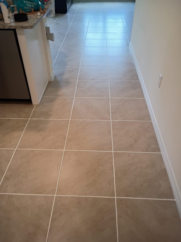 Tan square tile floor in a kitchen hallway with white grout, leading toward a lighter tiled area near an appliance.