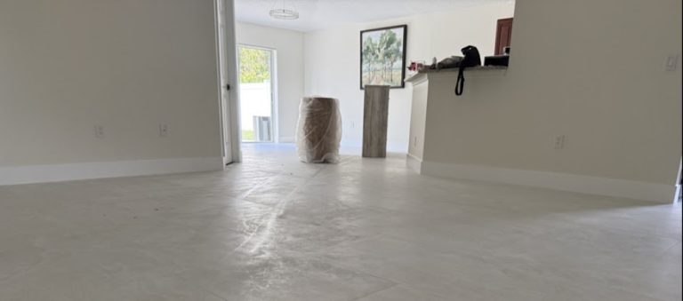 Empty room with light grey polished tile flooring, white walls, and a wrapped item on the floor near a decorative pedestal and framed painting.